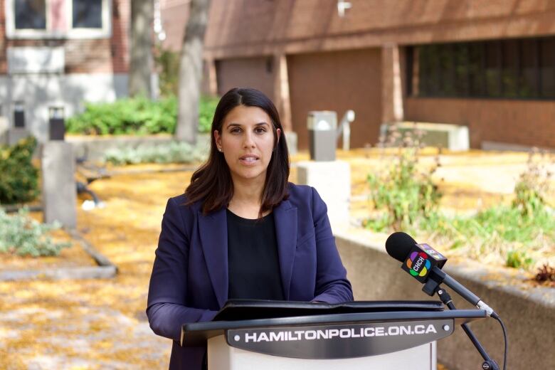 A woman in a blazer speaks outdoors at a podium with 