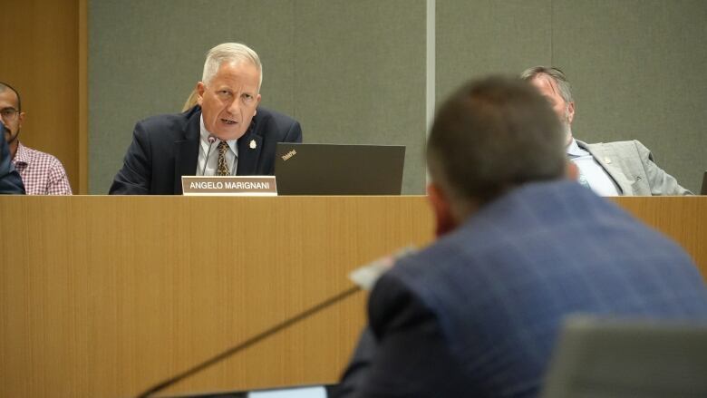 A man sitting down in an elevated council chamber asks questions of another man sitting at a microphone. 