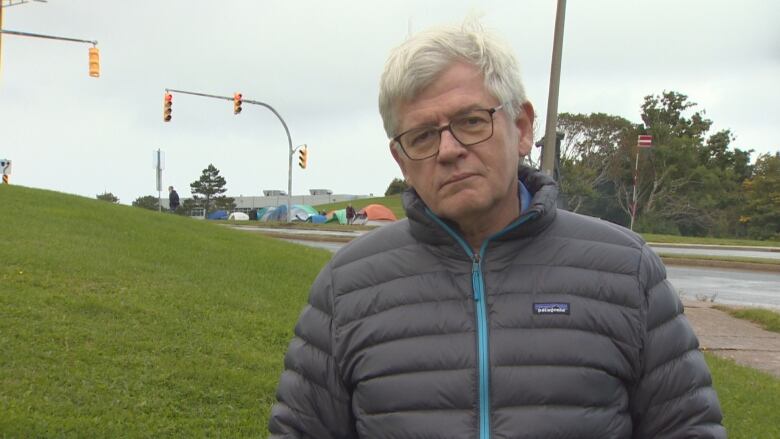 A man wearing a grey coat stands on a lawn. Several tents pitched across the street are seen over his right shoulder.