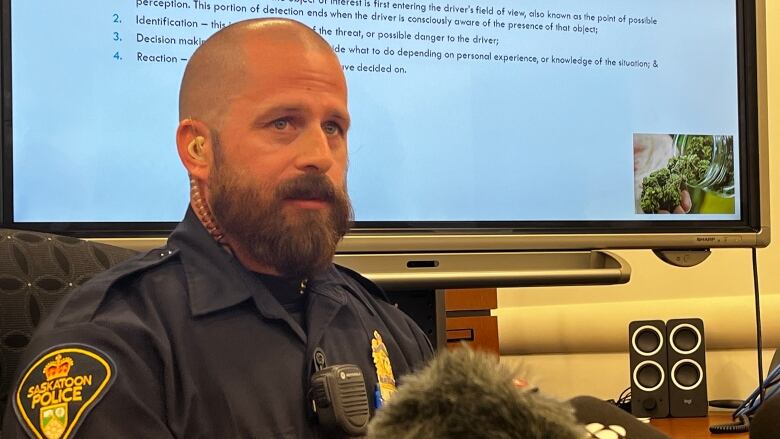 A man in a police uniform can be seen sitting in front of a screen with multiple news microphones in front of him. 