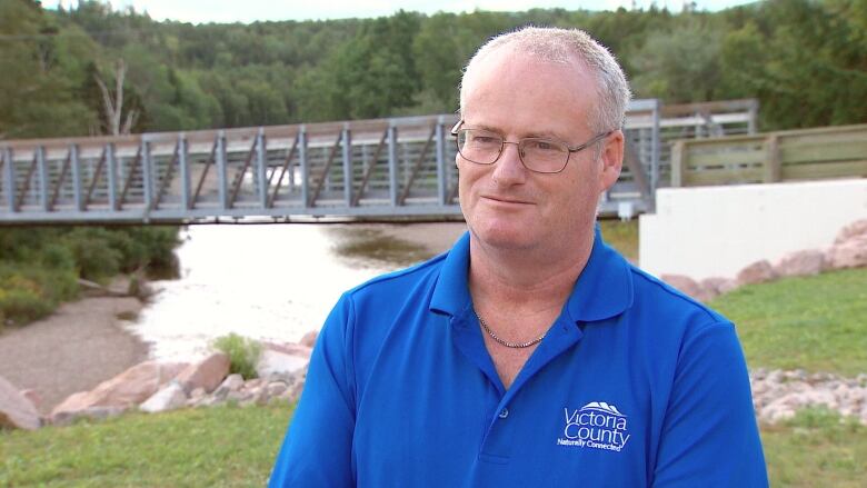 A man stands in front of a brrok with a bridge over it, with grey hair and glasses and wearing a blue shirt with a Victoria County logo on it.