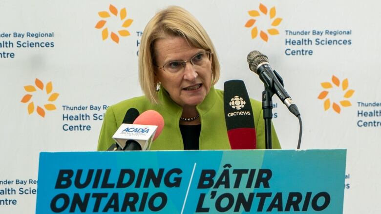 A woman with a blond bob and glasses leans into microphones on a podium in front of a backdrop that reads 