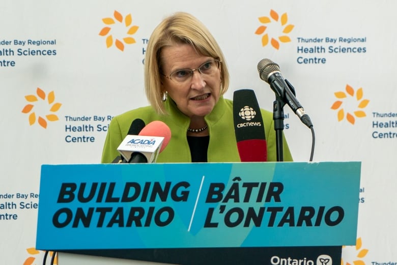 A woman with a blond bob and glasses leans into microphones on a podium in front of a backdrop that reads 