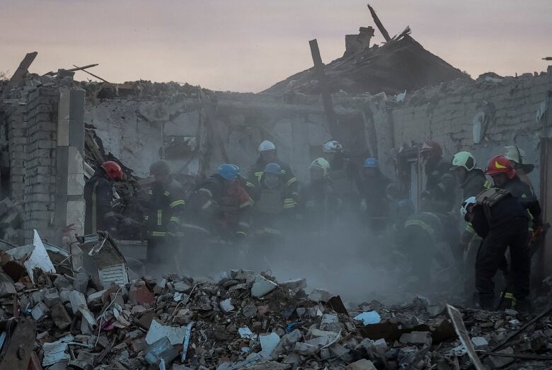 Several helmeted first responders are shown walking in the rubble of a building in which the roof has been blown off.