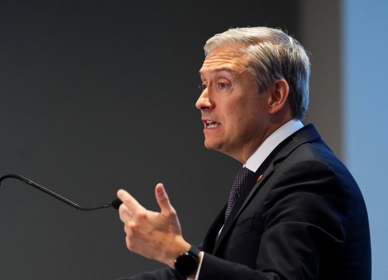 A man in a suit gestures with his left hand as he speaks into a microphone at a podium.