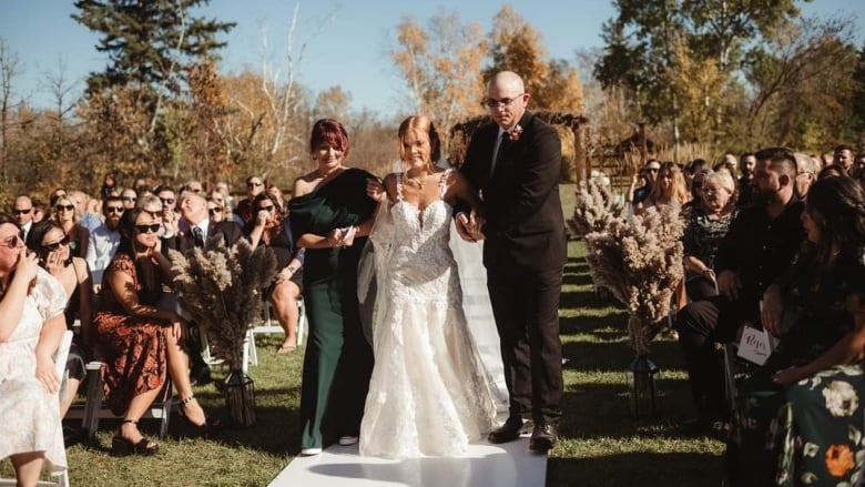 A woman in a white wedding dress walks down the aisle on her wedding day. On either side, her parents support her.