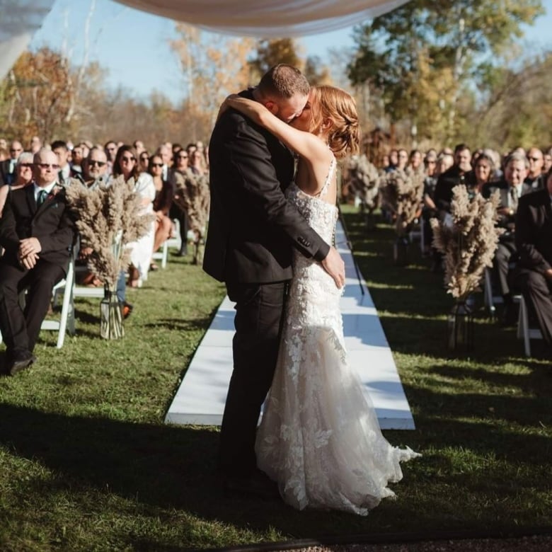 A woman in a white dress and a man in a black suit stand at a wedding altar at an outdoor wedding, as their guests watch.