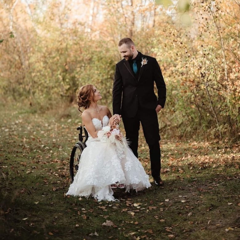 A woman in a white wedding dress, sitting in a wheelchair, holds hands with and looks up at her new husband - standing beside her wearing a black suit.