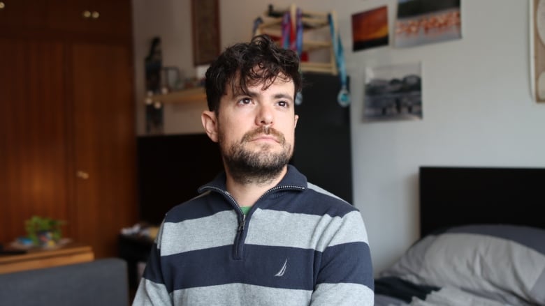 A man wearing a grey striped sweater is sitting on his bed looking outside the window. 