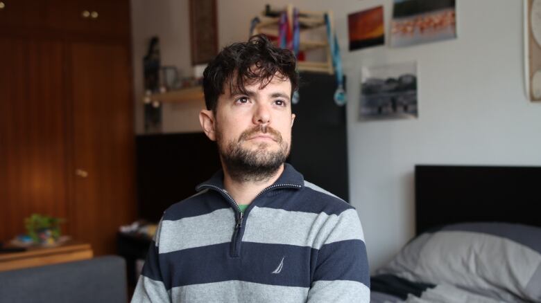 A man wearing a grey striped sweater is sitting on his bed looking outside the window. 