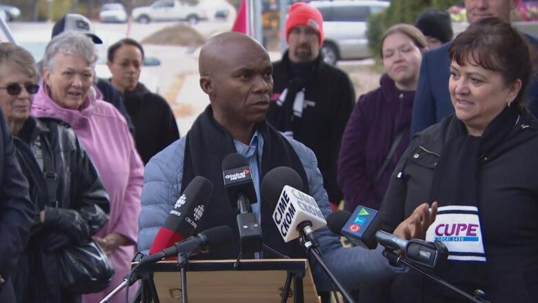 A man wearing a blue jacket and a black scarf can be seen talking at the podium with people around him.