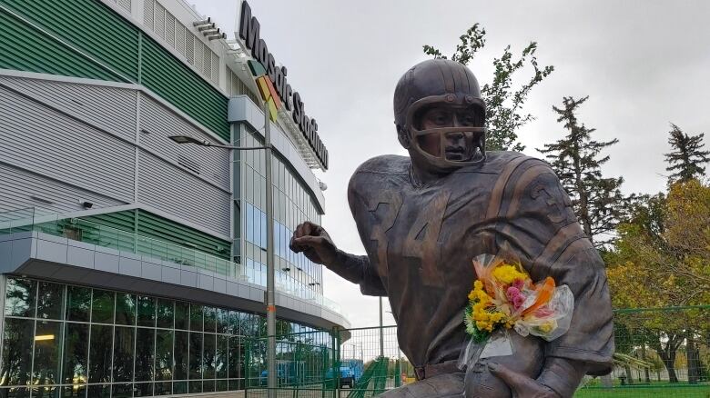 A bronze statue of a football player wearing number 34 and carrying a football stands in front of a part and a football stadium. A bouquet of colourful flowers is tucked into the statue, between the player's arm and the ball.