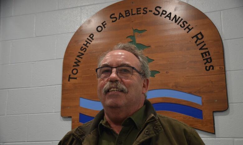 A man with a mustache stands in front of a sign that reads 'Township of Sables-Spanish Rivers'