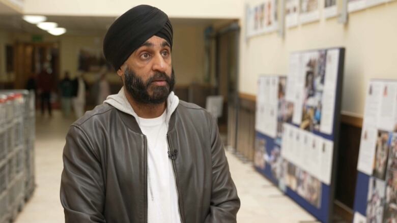 A man wearing a white hooded sweatshirt, a leather bomber jacket and a black turban speaks while standing in a building's lobby.