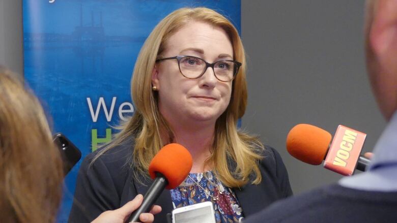 A woman with strawberry blonde hair and glasses standing in front of microphones.