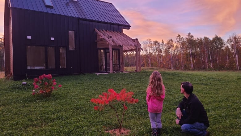 A man kneels on a lawn next to a girl. They both look at a home set against a pink sunrise. 