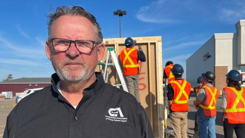 A man wearing glasses and a black button up shirt stands against a backdrop of apprentice workers practicing on a structure.