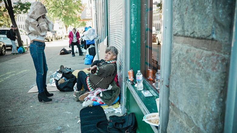 A senior woman with short grey hair sits outside of a closed storefront on a sidewalk, among piles of her belongings, which includes clothing.