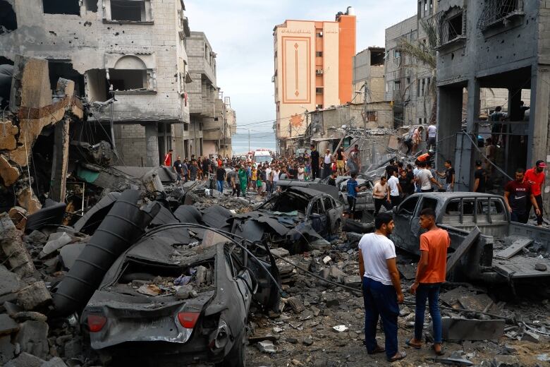 People stand in a narrow urban street that is filled with rubble and burned out cars.