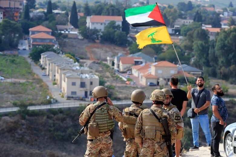 Soldiers on cliff next to man with yellow Hezbollah flag that contains a green machine gun