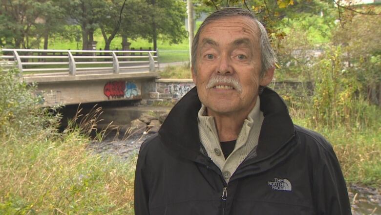 An man with grey hair and a mustache stands on an urban walking trail next to a river.