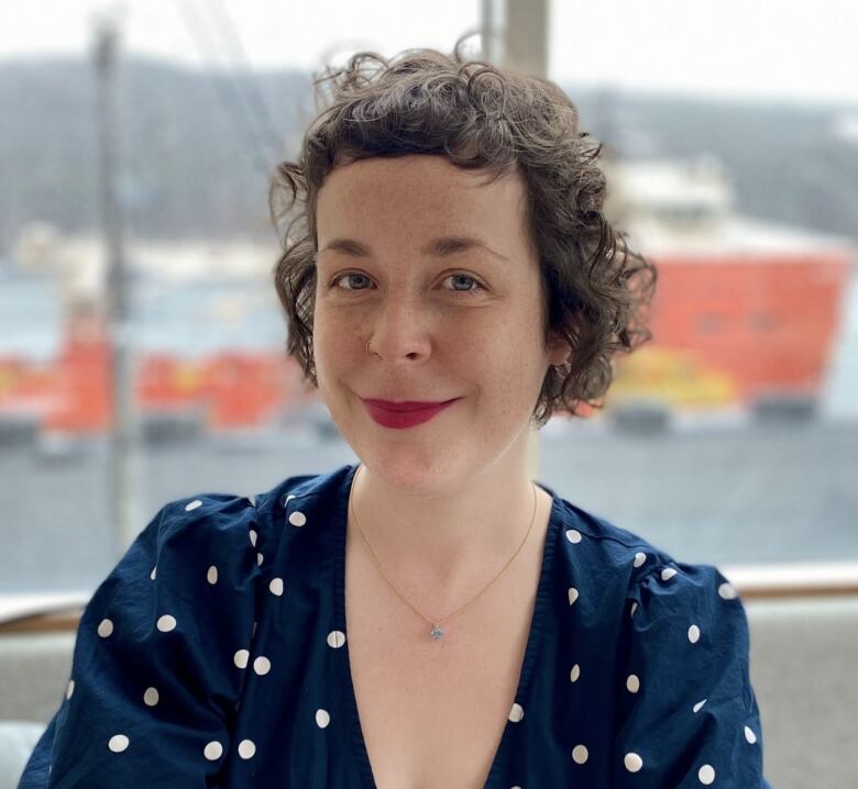 Woman with short, curly, brown hair, with a closed-mouthed smile and red lipstick in white polka-dot navy blue shirt. In the background through a window is a blurred image of a boat.