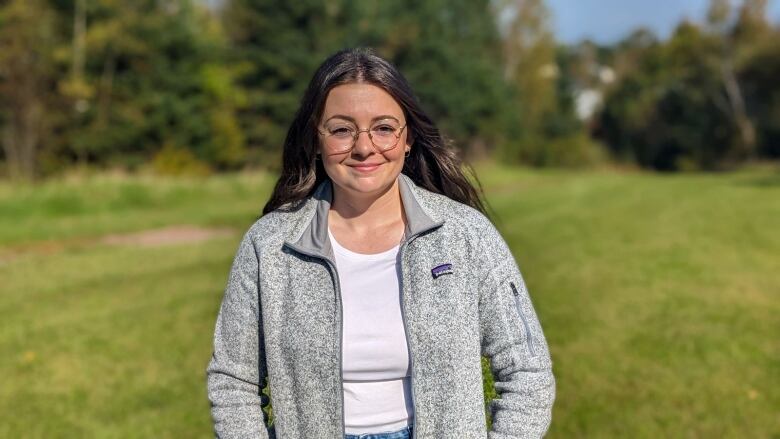 A woman with long brown hair and glasses stands in a treed area.