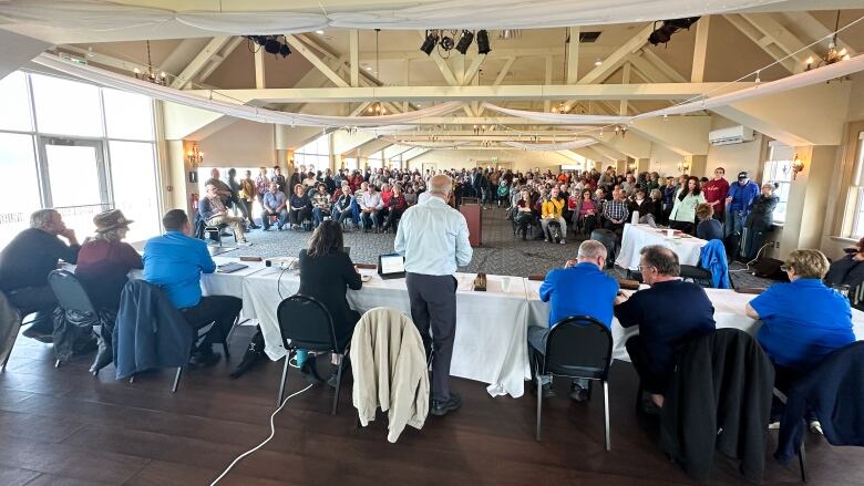 Eight people have their backs to the camera facing a large crowd of people standing and sitting in a large room. 