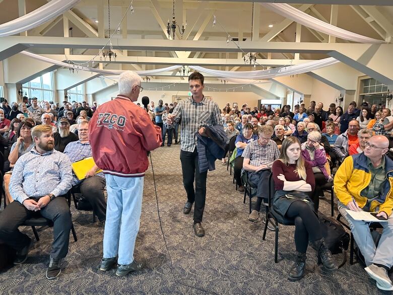 A man in a plaid shirt approaches another man with a microphone in the middle of a crowded room.