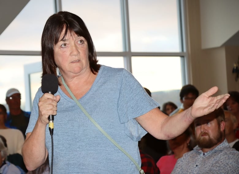 A woman in a blue blouse with dark hair speaks into a microphone and gestures with her hand.