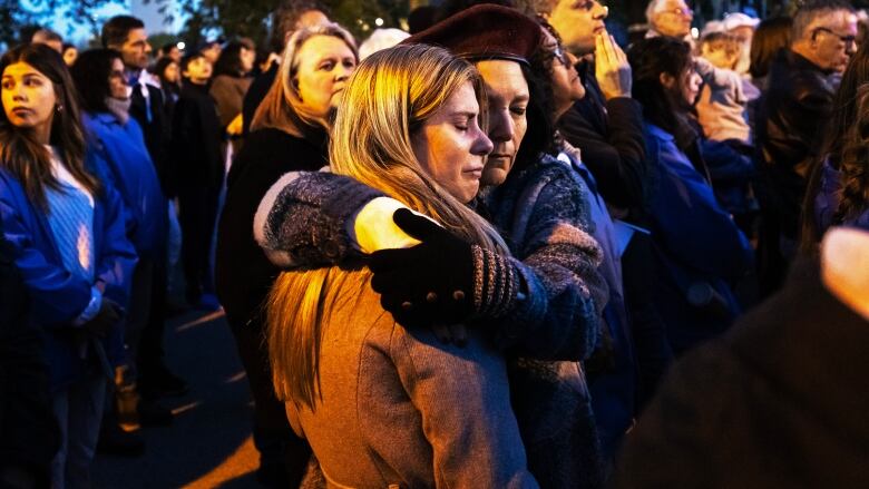 One woman hugs another who is weeping as they stand in a crowd of people outside under the evening sky.