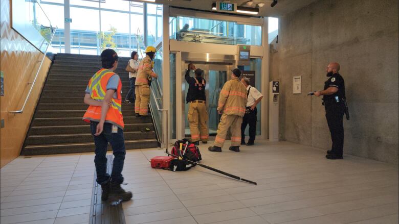 Firefighters in uniform work on a glass-doored elevators while a few people look on.