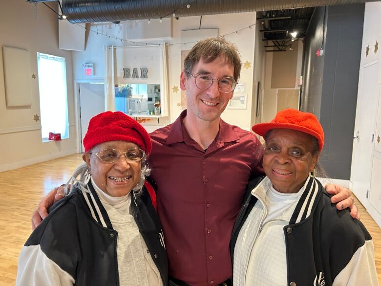 A man stands with his arms around two older Black women, wearing matching varsity jackets and red hats. They smile at the camera. 