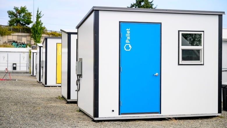 A small white structure with a blue door sits at the front of a row of a identical structures, one of which has a yellow door.