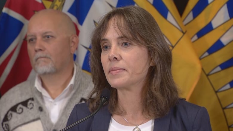 A white woman and an Indigenous man speak in front of a B.C. flag.