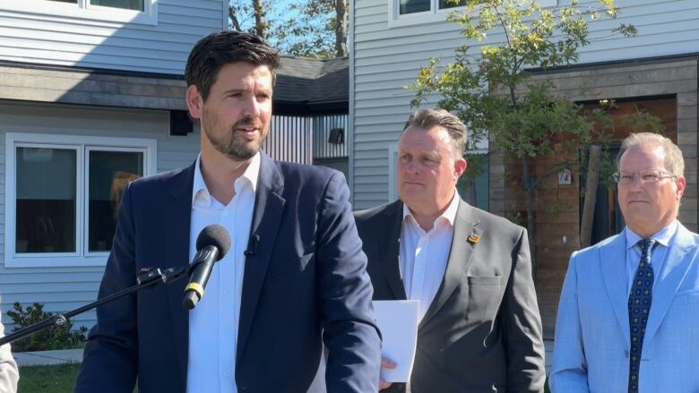 Three men wearing suits stand outside a residential building in front of a podium with a microphone.