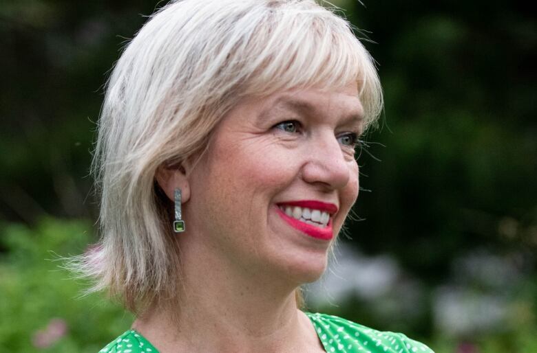 A smiling woman with short blond hair, wearing a green dress with small white dots, looks to her left.