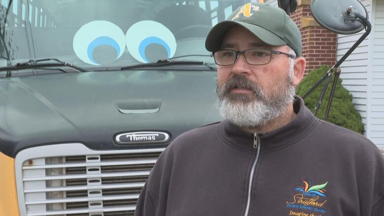 A man with a grey baseball cap and a black zip up jacket stands in front of his school bus.