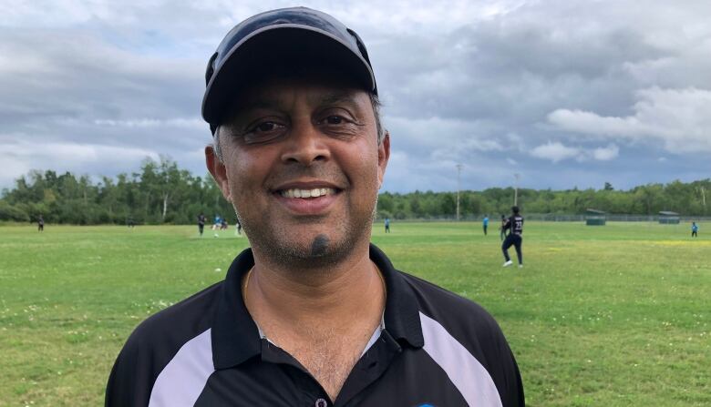 A smiling man with a ball cap standing in front of a grassy sports field.