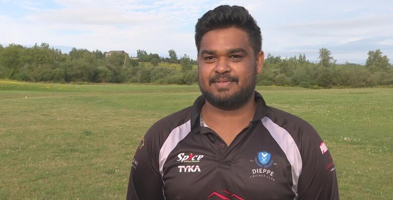 A smiling man wearing a long-sleeved sports jersey stands in front of a grassy sports field.