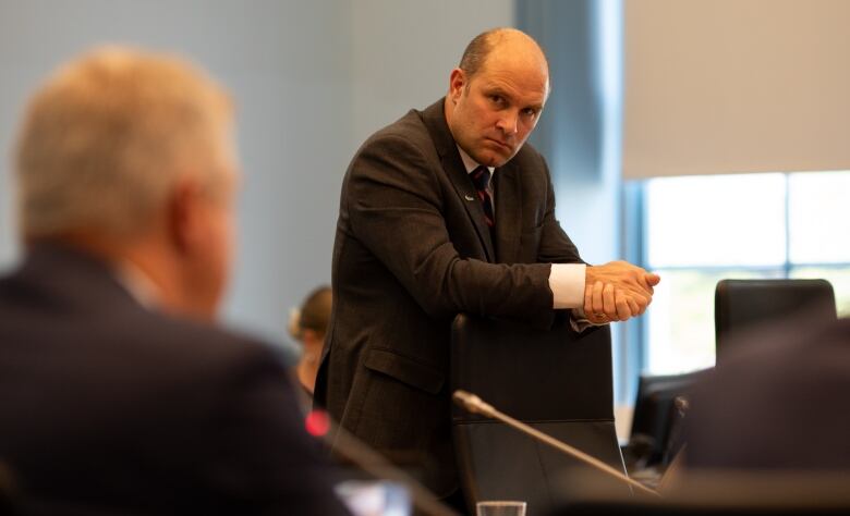 A city councillor leans on a chair during a meeting, listening to a speaker.