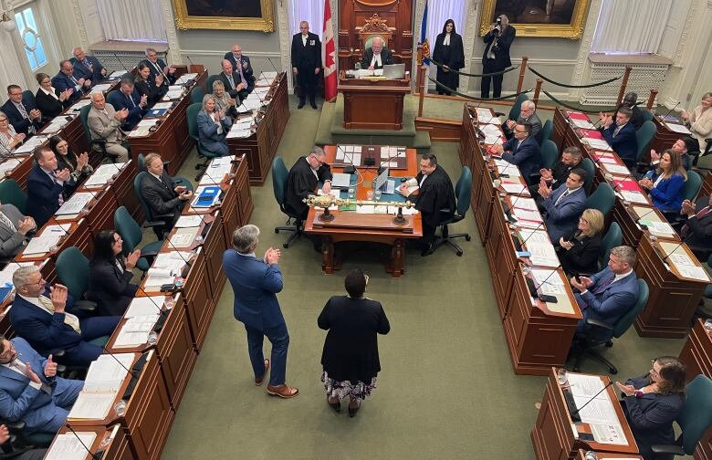 Two people stand in a room full of people sitting.