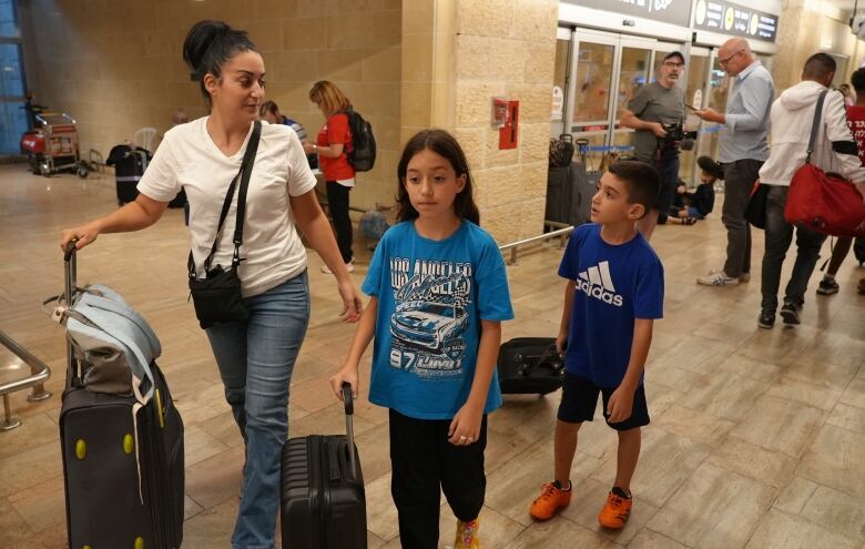 A woman and two children wheel luggage through a crowded airport concourse. 