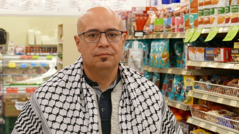 A man, wearing glasses and a black and white shawl draped over his shoulders, stands in front of a grocery store shelf lined with food.