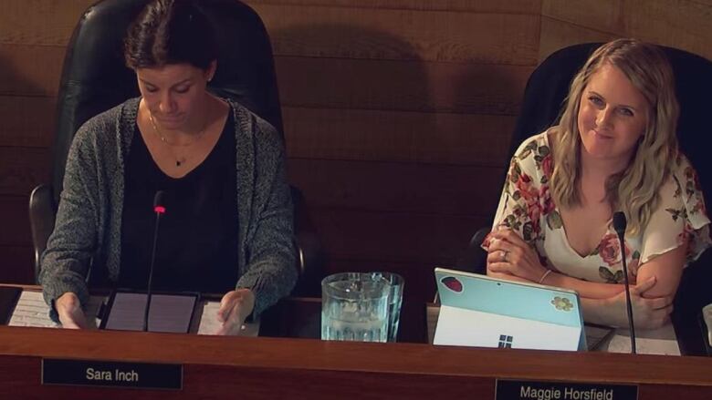 A woman with dark hair looks down, while a woman with blonde hair smirks 