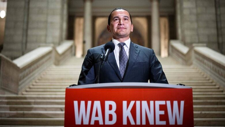 A man in black hair pulled back into a braid, stands at a podium. The name Wab Kinew is written on a card hanging at the front of the podium.