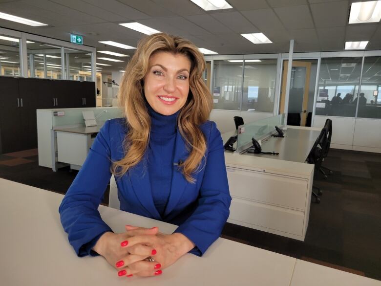 A woman in a blue turtleneck and blazer leans on a ledge with an office space in the background