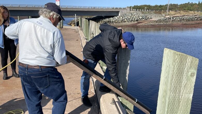 Two men lower a large metal rod into the water from a wharf. 