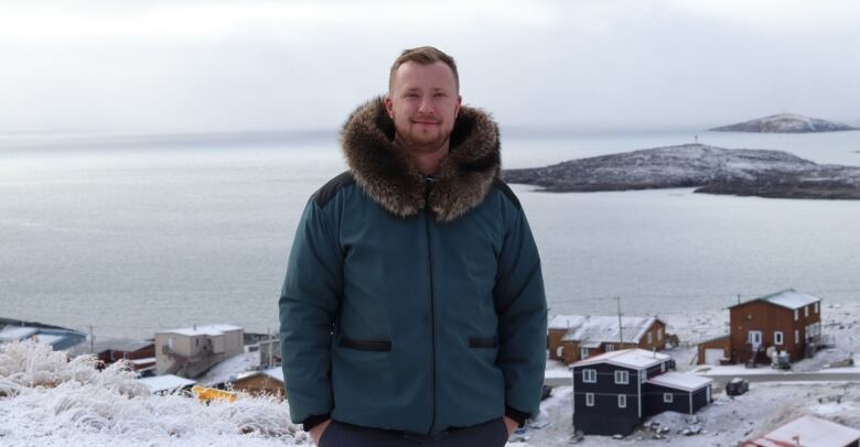 A man wearing a blue parka with dark brown fur poses for a photo with Iqaluit in the background. 