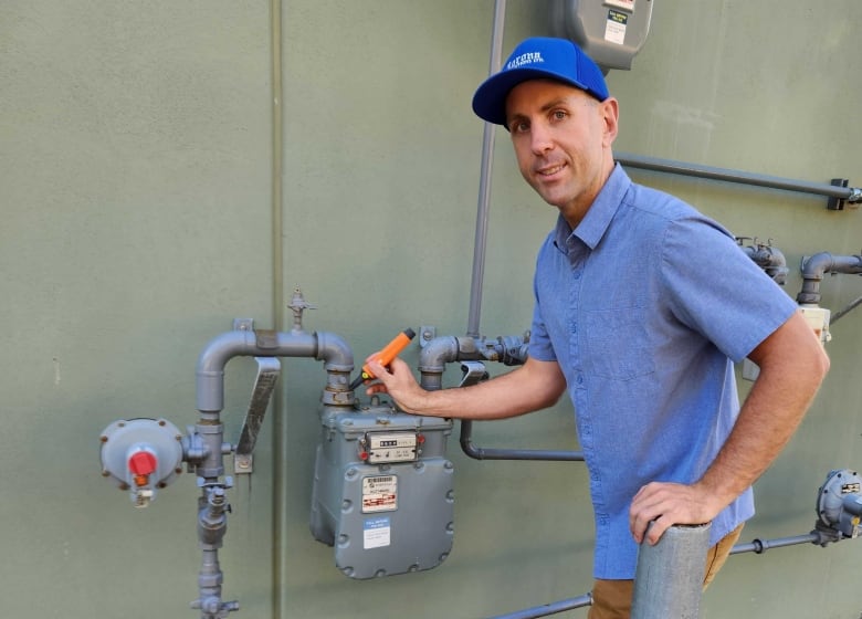 A man in a blue shirt and blue baseball hat stands beside a gas meter holding an orange device that's used to detect gas leaks. 